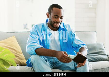 Positive black man in casual putting money in wallet and smiling, sitting on couch in living room interior Stock Photo