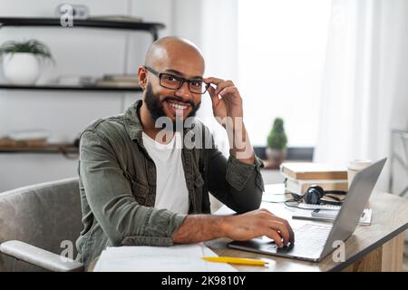Portrait of positive latino male freelancer working on laptop and smiling at camera, sitting at desk at home, free space Stock Photo