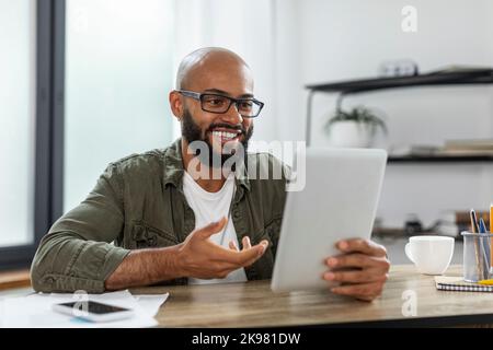 Video conference. Cheerful latino man having web call using digital tablet sitting at desk, talking at webcam in home Stock Photo