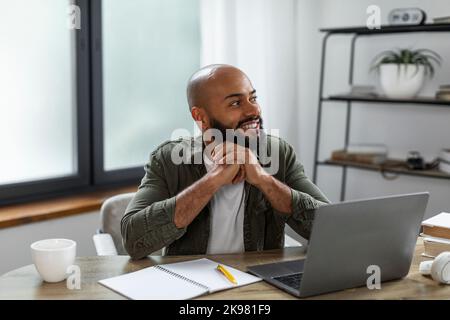 Happy latino man sitting at table in front of laptop computer at home, looking aside and thinking, copy space Stock Photo