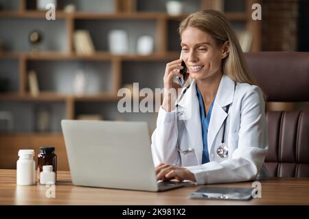 Smiling Doctor Woman Talking On Cellphone And Using Laptop At Workplace Stock Photo