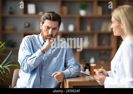 Sick Young Man Coughing During Meeting With Female Doctor In Clinic Stock Photo