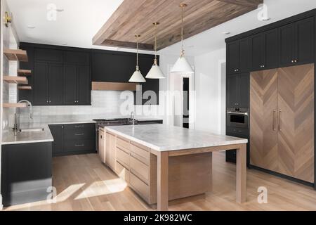 A luxury black kitchen with a pendant lights and wood frame over a large island, stainless steel appliances, and a wood paneled refrigerator. Stock Photo
