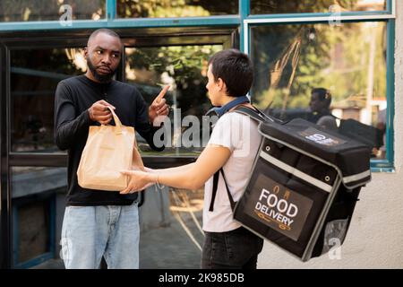 Unsatisfied client refuse taking food order, bad delivery service, courier and angry customer. Woman giving upset employee takeaway meal near office building outdoors, medium shot Stock Photo