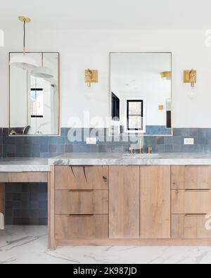 A luxury home's renovated bathroom with blue tiles, a light wood vanity cabinet, gold lights mounted on the wall, and a grey stone counter top. Stock Photo
