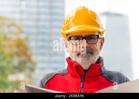 Portrait of a confident, caucasian, bearded, grey-haired, middle-aged professional construction worker in uniform wearing a bright orange helmet and eyeglasses, holding a folder with documents, looking at the camera. High quality photo Stock Photo