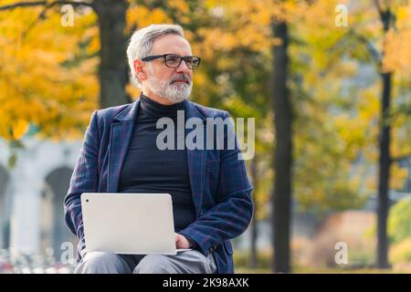 Modern stylish bearded grey-haired mature man entrepreneur sitting on a bench in the park with fall foliage and working on his laptop, checking business emails. High quality photo Stock Photo