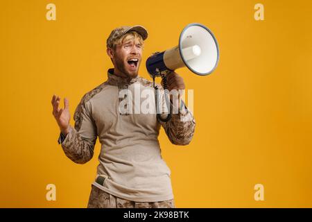European military man wearing uniform shouting in megaphone isolated over yellow background Stock Photo