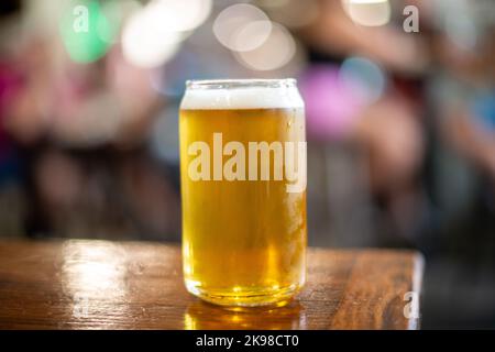 A clear drinking glass in the shape of a beer can filled with cold froth lager ale. The Belgian pint sits on the edge of a wooden patio table. Stock Photo