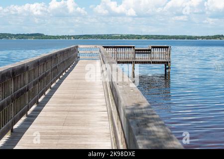 A long wooden finger pier wharf with wood railings stretching out into the blue tranquil water. There's land in the distance with a white cloudy sky. Stock Photo