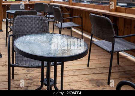 Multiple empty black metal tables with round glass tops and black rattan chairs on a sidewalk cafe patio of an outdoor restaurant. Stock Photo