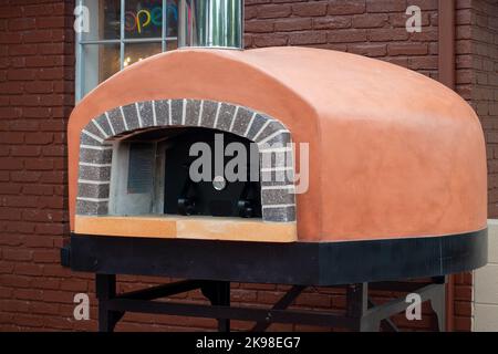 An exterior of a gas clay pizza oven on black metal legs on a restaurant patio. There are bricks surrounding the opening of the Italian equipment. Stock Photo