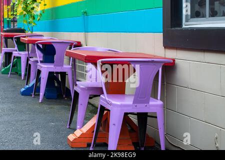 An outdoor restaurant or cafe eating area with bright yellow, green and blue stipes on the exterior wall. The patio tables are colorful red metal Stock Photo