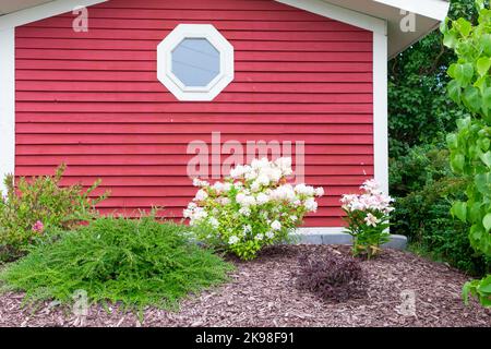 A red wooden shed with an octogen shaped glass window in the middle of the wall. The trim on the building is white. There are shrubs and flowers in fr Stock Photo