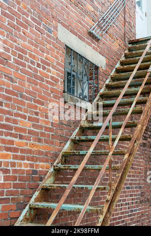 The exterior corner wall of an old brown and red colored brick building with rusty metal fire escape stairs. There's a multi-pane glass window Stock Photo