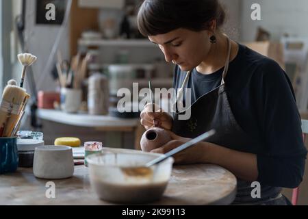 Young girl entrepreneur sits at table in ceramic workshop and applies paint to homemade vessel Stock Photo