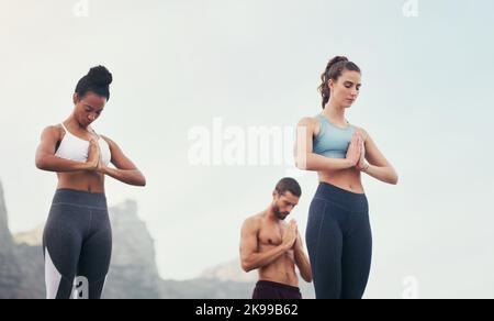 Meditation is the best medication. a group of young people standing and meditating together while on the beach during an overcast day. Stock Photo