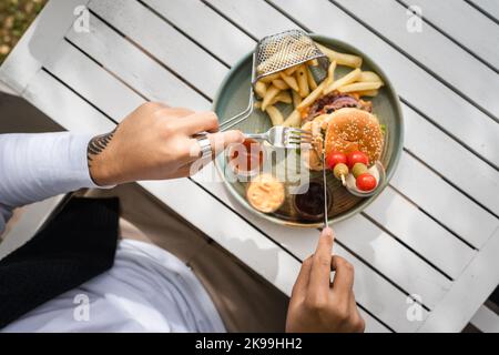 close up on burger in a plate and hands of unknown caucasian man prepare to eat on the table in sunny day outdoor at restaurant hold fork and knife Stock Photo