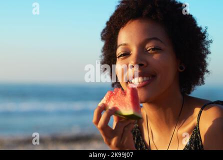 The best summer fruit for days like these. Portrait of an attractive single young woman eating a watermelon piece on the beach during the day. Stock Photo
