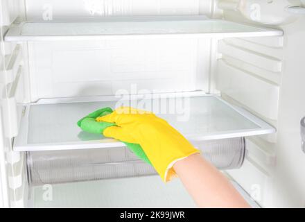 Hand cleaning refrigerator. Person washing refrigerator with rag. Housekeeper wipes shelves of clean refrigerator. Hand in yellow rubber protective gl Stock Photo