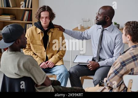 Young man talking about his problems with psychiatrist supporting him at psychology class with other young people Stock Photo