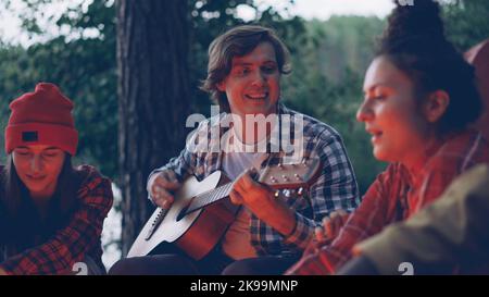 Handsome young man tourist is playing the guitar and smiling while his happy friends are singing and having fun resting around fire near lake or river. Green trees are visible. Stock Photo