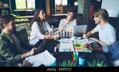 Multi-ethnic group of colleagues is discussing charts and graphs sitting at desk in modern office during team meating and sharing ideas. Young people, teamwork and workplace concept. Stock Photo