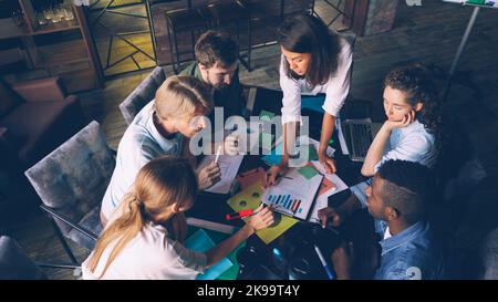 Multiracial workgroup is analyzing diagram and writing on colored stickers sitting at desk in office room. High angle view of people talking, pointing at charts and taking notes. Stock Photo