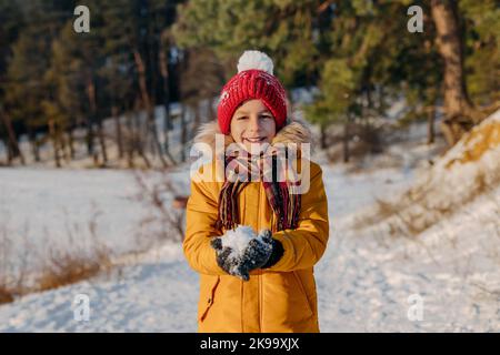 Happy kid holding in hands snowball on the background of snowy forest. Smiling boy in warm clothing having fun outdoors. Concept of christmas holidays Stock Photo