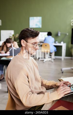 Serious businessman in eyeglasses concentrating on his online work on digital tablet at table at office Stock Photo