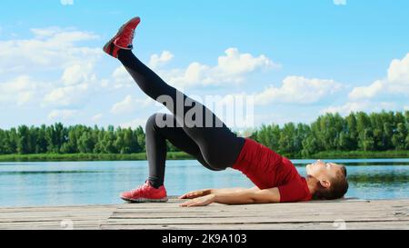 Beautiful, athletic young blond woman stretching, doing different exercises, jumping, lunges, squats. Lake, river, blue sky and forest in the background, summer sunny day. High quality photo Stock Photo