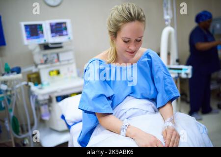 Taking it all in before I go under the needle. a young pregnant woman sitting on her hospital bed and looking thoughtful with a nurse in the Stock Photo