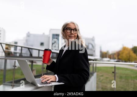 Middle aged older business woman works on a laptop and drinks coffee on the office terrace, concept of successful business development Stock Photo