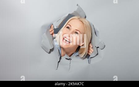 Charismatic joyful woman with blonde hairstyle, looking playfully, woman dreaming, looking up, stands in torn paper hole, notices funny pleasant. Gray Stock Photo