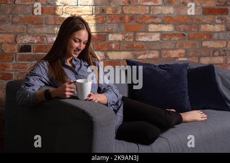 Woman in a blue shirt lying down on the couch and looking into a white cup Stock Photo
