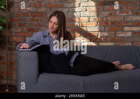 Woman in a blue shirt lying on the couch and writing in a notebook with a pen Stock Photo