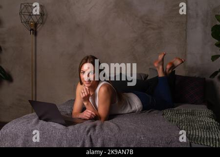 Smiling woman in jeans and a white top lying on her stomach in bed with a laptop in front of her Stock Photo