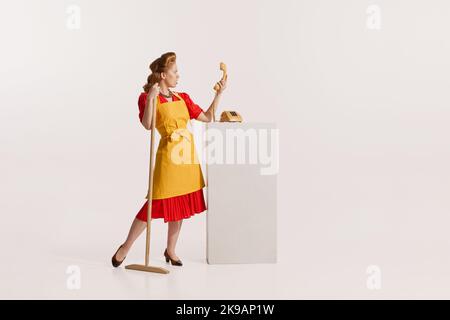 Portrait of elegant redhead woman with beautiful hairstyle cleaning with mop and talking on vintage phone isolated over white background Stock Photo