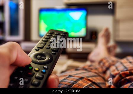 Person lying on bed with crossed legs and switching television channels on TV with finger on remote controller Stock Photo
