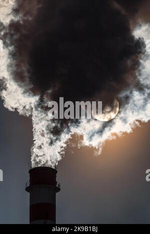 factory chimney and coming out dark smokes in the sky and sun at background. Stock Photo