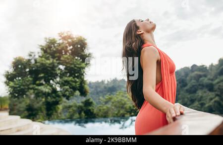 Side view shot of relaxed young woman standing at a railing. Female in orange dress at poolside in luxury resort. She is enjoying the climate and fres Stock Photo