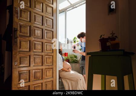 young latin caucasian man with a beard is standing drinking mate talking to his pregnant wife at home, seen through the front door from inside the hou Stock Photo