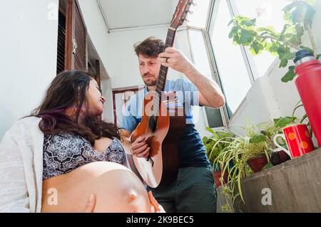 Pregnant young Latin couple of Argentine Caucasian man and Brazilian woman, they are at home enjoying, singing songs with the guitar and drinking mate Stock Photo