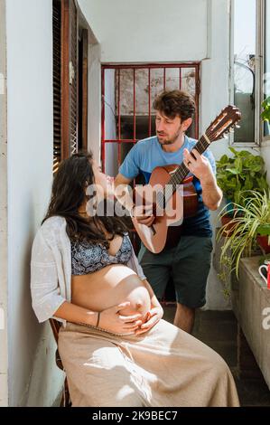 latin caucasian young man with beard standing at home, sings and plays a song with his acoustic guitar to his wife who is pregnant and sitting watchin Stock Photo