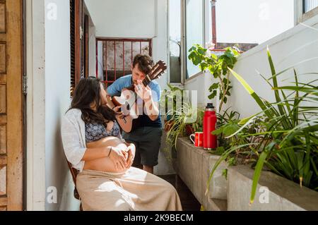 young latin couple of bearded caucasian argentinian man singing and playing guitar with his pregnant brazilian woman wife, at home Stock Photo