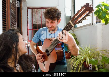 young latin caucasian man with a beard, at home smiling serenades him with his guitar and sings songs to his pregnant wife who is happy to see him. Stock Photo