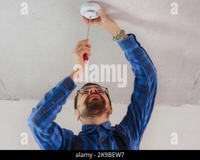 young electrician man in blue work suit installing smoke detector on the ceiling Stock Photo