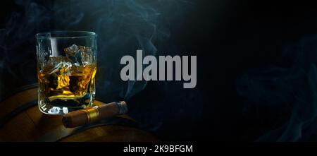 A glass of whiskey with ice on an oak barrel and a smoking Cuban cigar on a dark background. Men's club banner idea. Copy space for text Stock Photo