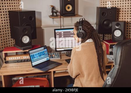 African American anchor woman sitting in front of microphone and laptop with mixing board in recording studio Stock Photo