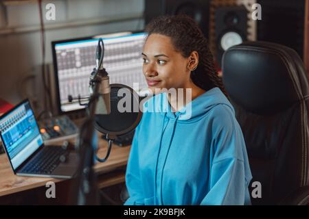 Charming ethnic woman, radio presenter with stylish dreadlocks working in a broadcasting audio recording studio Stock Photo